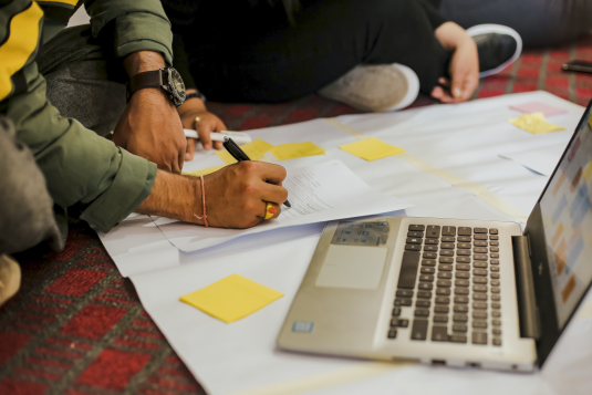 A photo of a man's hand writing on a piece of paper with a laptop by his side.