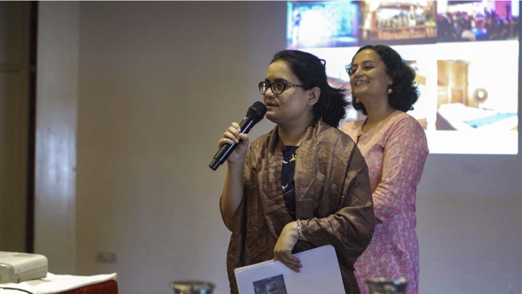 A photo of two women speaking to a crowd, standing in front of a presentation and holding a microphone.