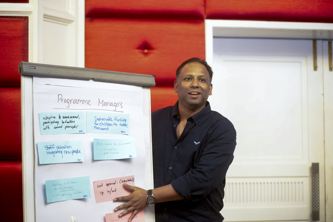 A photo of a man standing next to a flipbaord with notes from research meeting, speaking to the wider room.